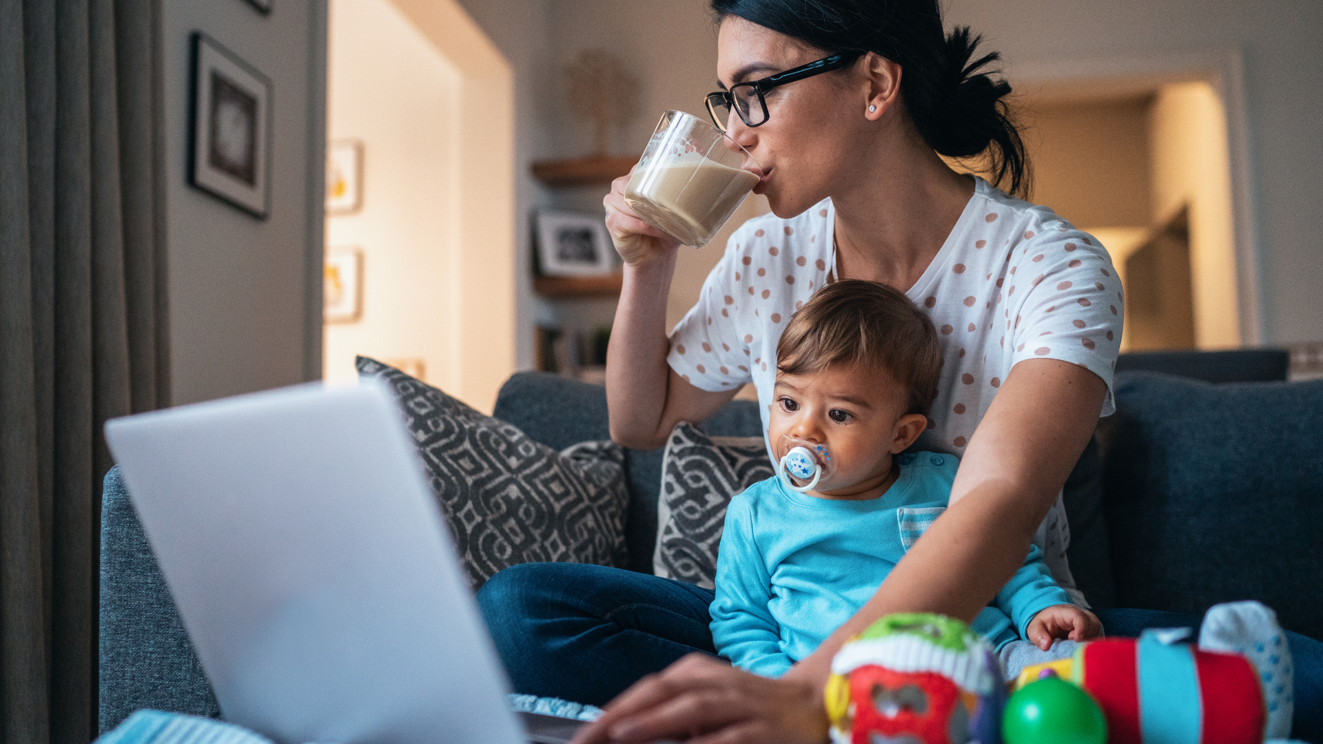 Frau sitzt auf dem Sofa mit einem Kleinkind auf dem Stoß. Sie bedient einen Laptop und trinkt dabei einen Milchkaffee. Das Kind schaut gebannt auf den Bildschirm.